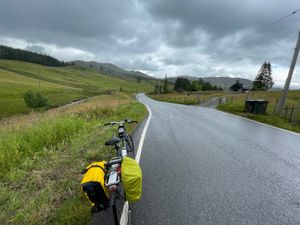 Red bike loaded with two pannier bags, looking out on a long, smooth asphalt road. Cloud-topped mountains loom in the distance.