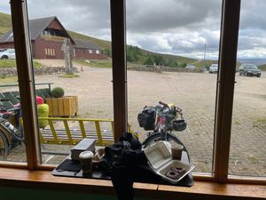 Inside a large rustic ski center, looking out the big window to the top of the Lecht. Bikes parked outside by a bench. Hot drinks and a container of two donuts sit on the windowsill.