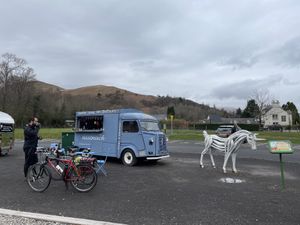 In the foreground, Jack stands by the bikes, drinking coffee at an outdoor table in front of a little retro blue coffee truck called 