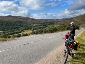 Jack takes a water break on a sunny day, looking back down a slight incline. His bike is parked in front of him, on the side of a two-lane road.