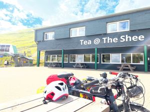 Two bikes lean against an outdoor picnic table, facing the large cafe at Glenshee Ski Centre on a blue-sky, sunny day. 